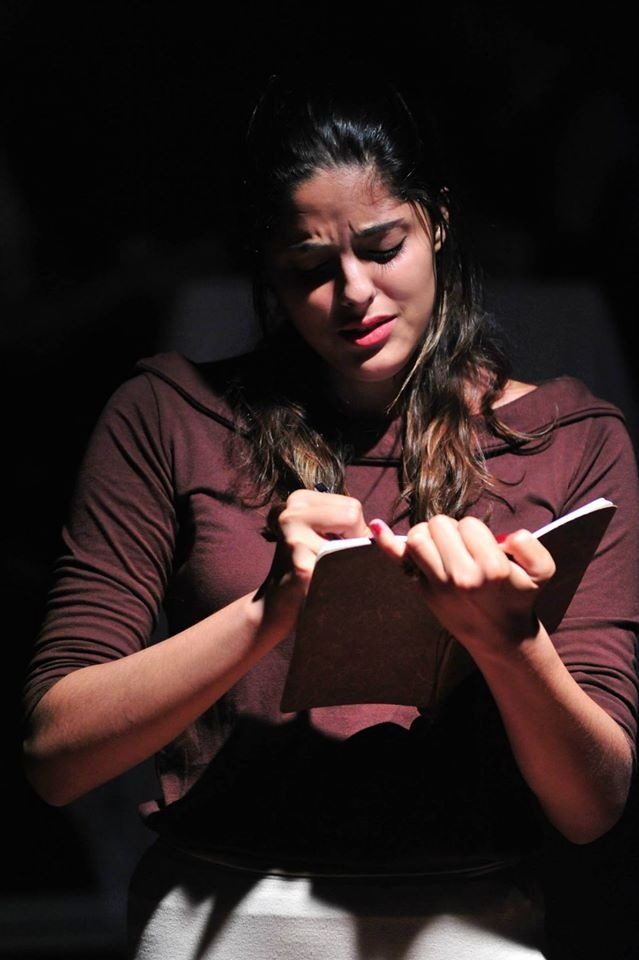 Amanda em cena de teatro, sob luz dramática, segurando um livro