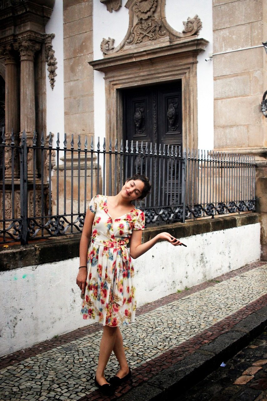 Amanda Cavalcanti em frente a uma igreja histórica, de vestido floral, sorrindo